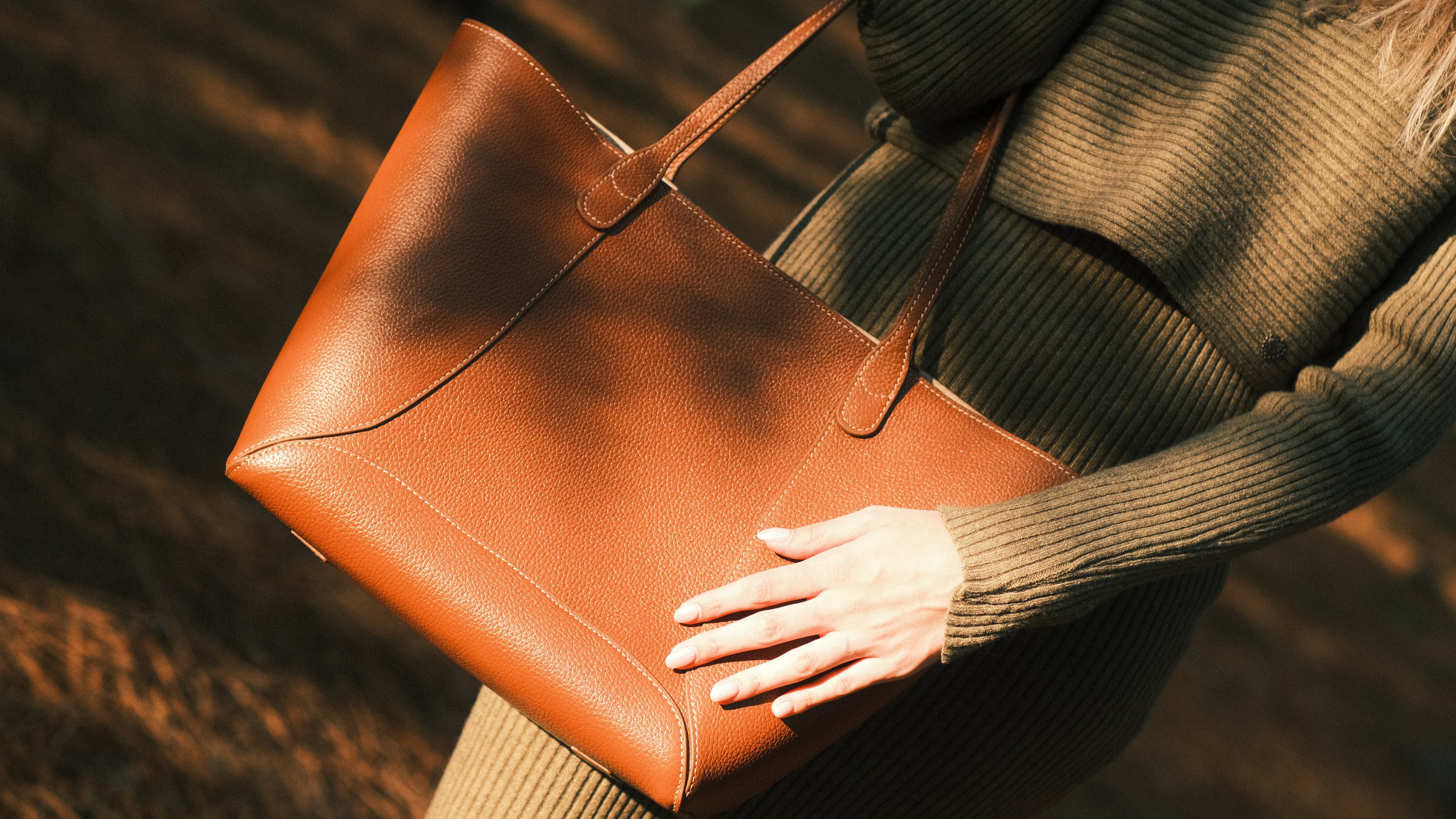 Brown leather tote bag held by a person wearing a green sweater.