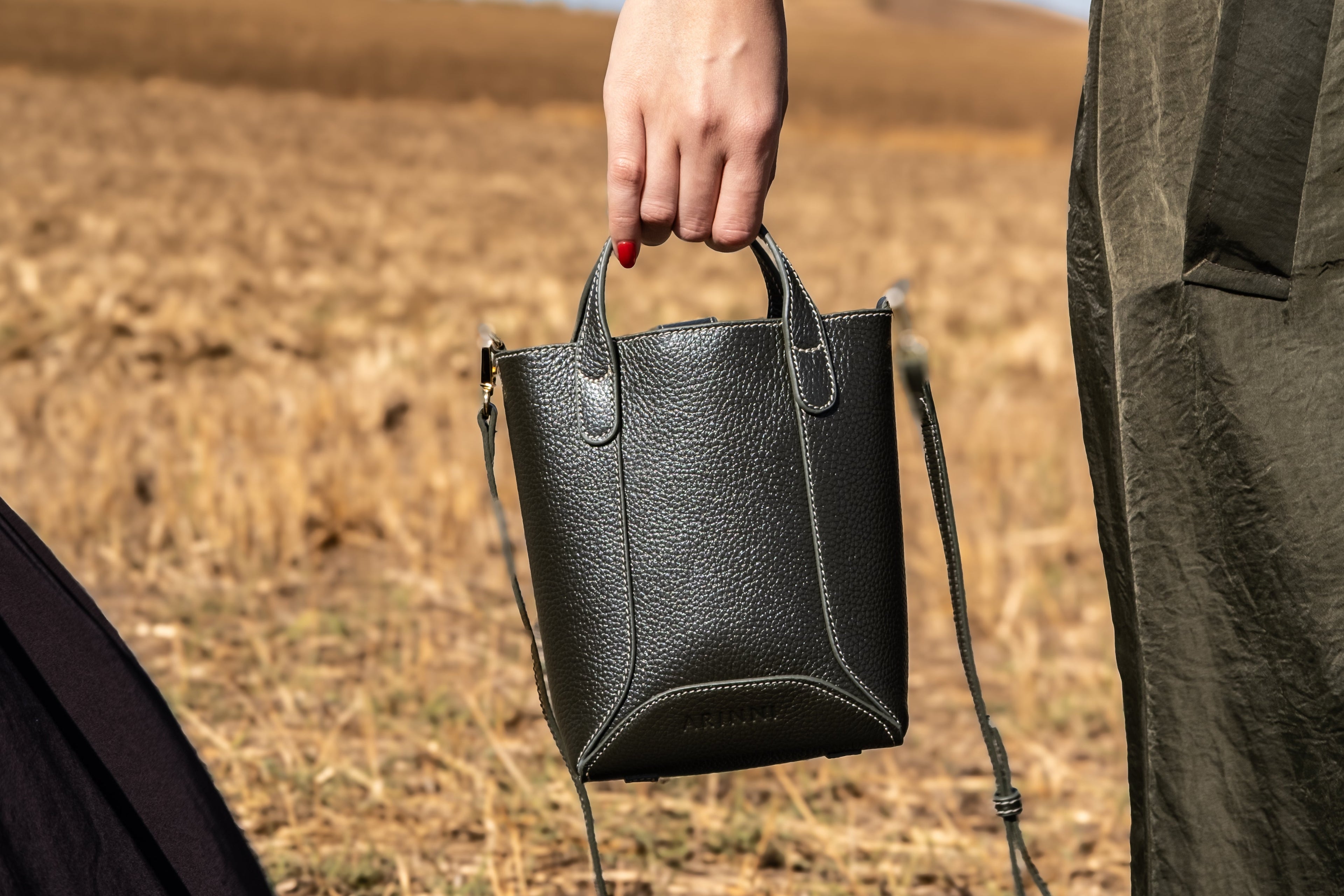 Person holding a dark green handbag in an outdoor setting with a blurred background