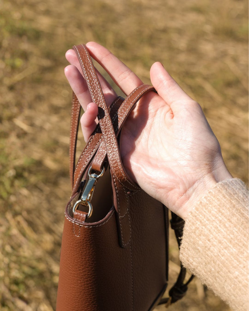 Brown leather bag held by a hand with a blurred grassy background