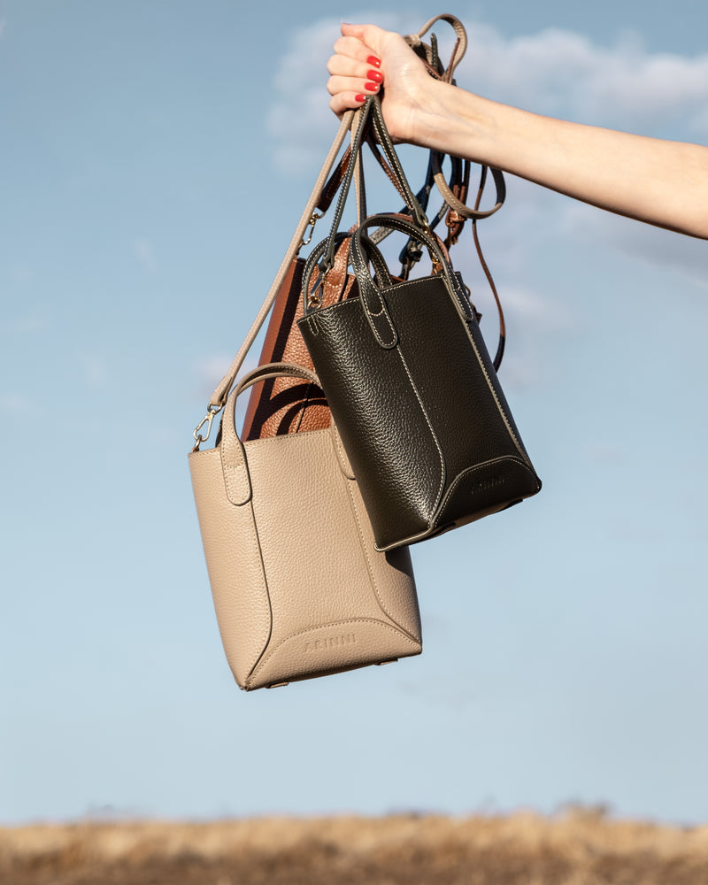 Three leather handbags, one beige, one dark green and one brown held against a clear blue sky.